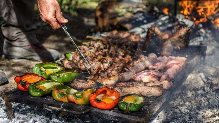 Auf einem Grillrost über Glut liegen bunte Paprikahälften, gefüllte Avocados und verschiedene Fleischstücke, daneben eine Grillzange.
