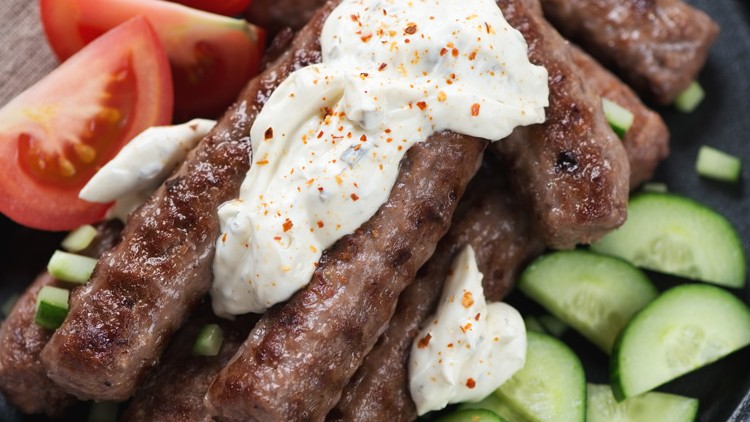 Gegrillte Hackfleischrollen mit weißem Dip, garniert mit Tomatenstücken und Gurkenscheiben auf einem dunklen Teller.