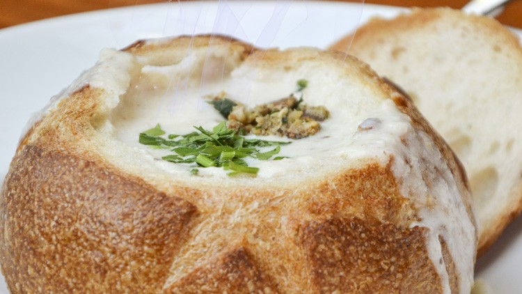 Ein runder Laib Brot dient als Suppenschale, gefüllt mit heller Suppe und garniert mit frischen Kräutern, daneben eine Brotscheibe.