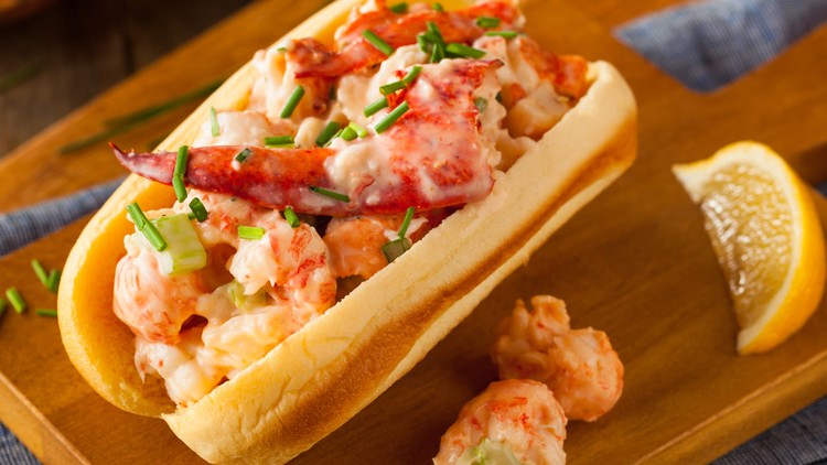 Ein Hotdog-Brötchen mit Hummer- und Garnelensalat, garniert mit Schnittlauch, liegt auf einem Holzbrett neben einer Zitronenscheibe.