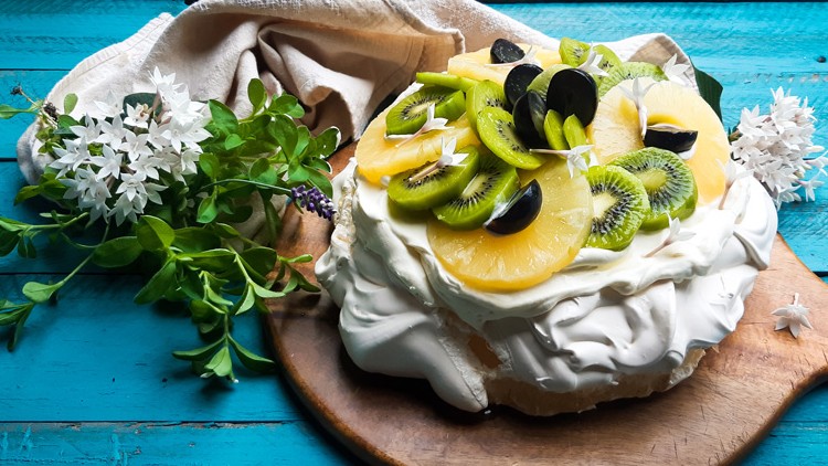 Eine Pavlova mit Sahne, Ananasscheiben, Kiwi und Trauben auf einem Holzbrett, dekoriert mit weißen Blüten und grünem Blattwerk.