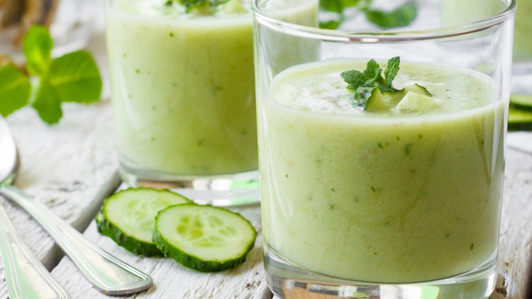 Zwei Gläser mit grüner Gurkensuppe, garniert mit Gurkenstücken und Minze, stehen auf einem hellen Holztisch mit Löffeln.