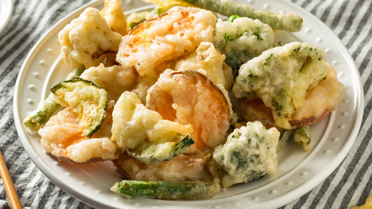 Krosser Tempura-Mix aus Garnelen und Gemüse wie Zucchini und Bohnen auf einem weißen Teller, daneben Sojasauce und Stäbchen.