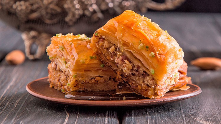 Zwei Baklava-Stücke mit goldbrauner Schicht aus Blätterteig und Nüssen liegen auf einem braunen Teller, im Hintergrund Dekoration.