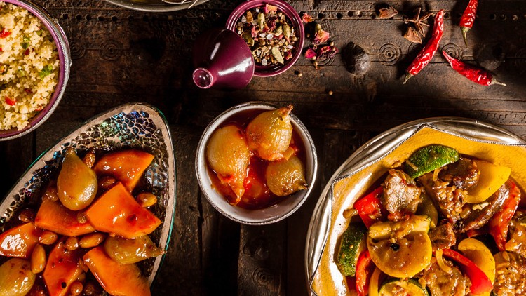Verschiedene Teller mit mariniertem Gemüse, Couscous und Gewürzen auf einem dunklen Holztisch, dekoriert mit roten Chilischoten.