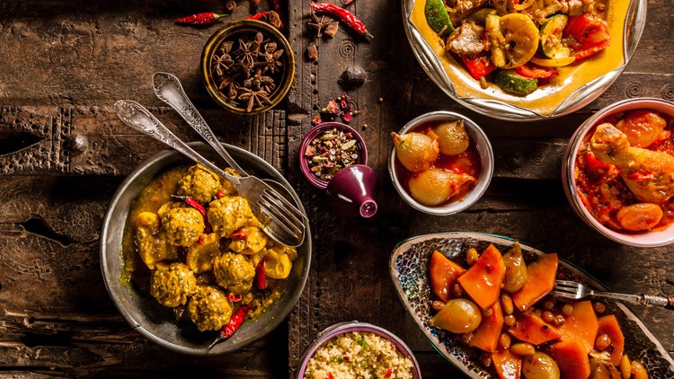 Mehrere Schalen mit orientalischen Speisen wie Gemüse, Fleisch und Couscous auf rustikalem Holztisch mit Gewürzen dekoriert.