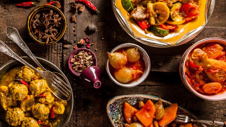Verschiedene orientalische Speisen wie Fleischbällchen, Geflügel und Gemüse in Schalen auf verziertem, dunklem Holztisch.