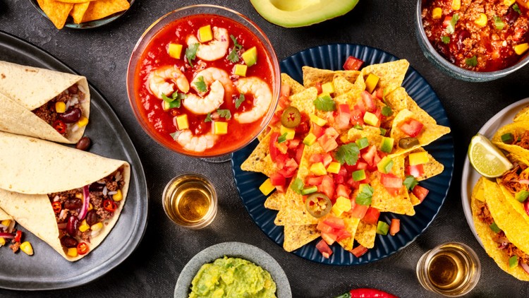 Verschiedene mexikanische Speisen wie Tacos, Tortillas, Nachos mit Gemüse, Guacamole, Garnelen-Cocktail und Limetten auf gedecktem Tisch.