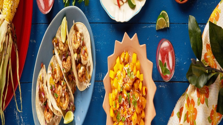 Gegrillte Garnelen-Tacos, eine Schale mit Mango-Salat, Maiskolben und Getränke auf blauem Holztisch mit farbiger Deko.