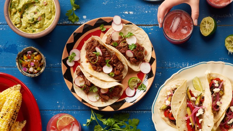 Verschiedene Tacos mit Fleisch oder Gemüse, Maiskolben, Guacamole und Gurkensticks auf einem blauen Holztisch.