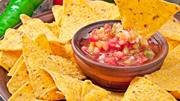 Gelbe Tortilla-Chips auf einem Teller mit roter, stückiger Salsa-Dip-Schale, dekoriert mit grüner Chili und Tomaten.