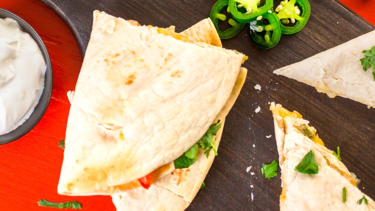 Aufgeschnittene Quesadillas mit Koriander, Jalapeños und einem Schälchen Sauerrahm auf einem dunklen Holzbrett.