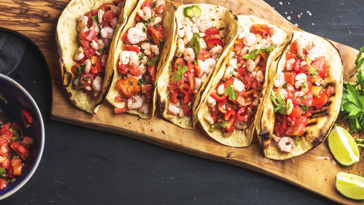 Fünf Tacos mit Garnelen, Tomatenstücken und Kräutern auf einem Holzbrett neben einer blauen Schüssel und Limettenspalten.