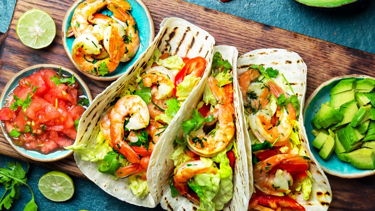 Drei gefüllte Tacos mit Garnelen, Salat und Gemüse auf Holzbrett, daneben Salsa, Limettenhälften und Avocadostücke in Schalen.
