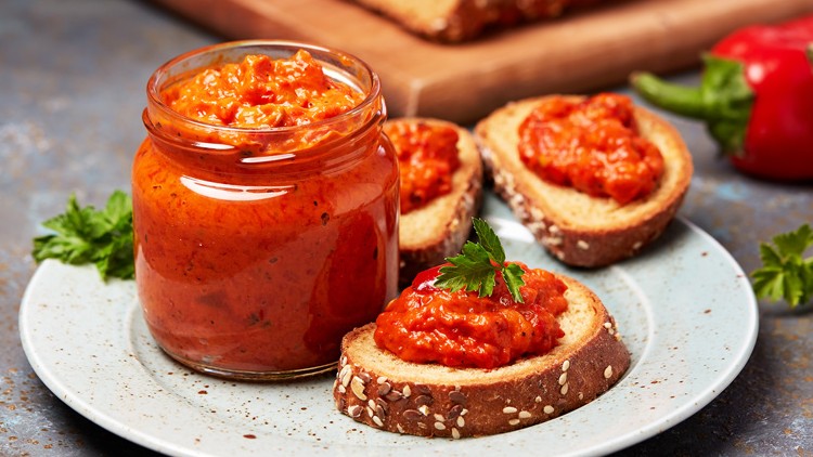 Ein Glas mit rotem Paprika-Aufstrich steht auf einem Teller mit drei belegten Brotscheiben, daneben liegen rote Paprikaschoten.