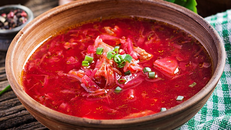 Rote Suppe mit Gemüse und frischen Kräutern in einer braunen Schüssel, daneben grün-weiß kariertes Tuch auf Holztisch.