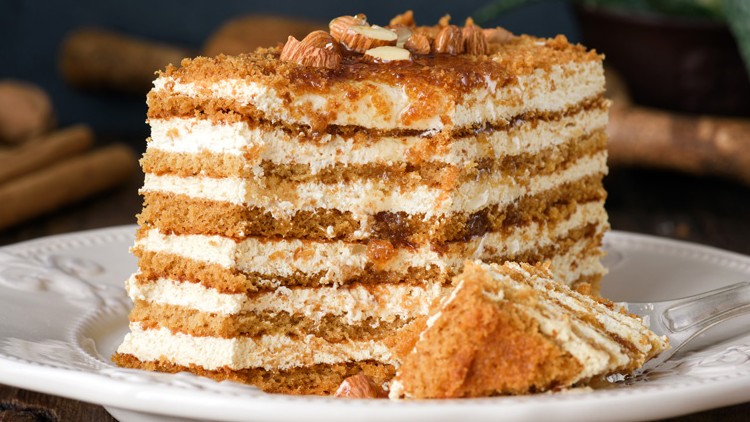 Stück mehrschichtige Honig-Torte mit Creme auf einem weißen Teller, garniert mit Nüssen, daneben eine Gabel.
