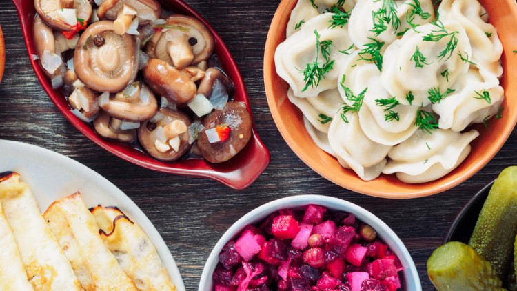 Verschiedene Speisen auf einem Tisch, darunter Pelmeni, gefülltes Brötchen, marinierte Pilze, Rote-Bete-Salat und Gewürzgurken.