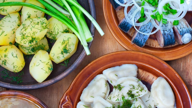 Gekochte Kartoffeln mit Dill, eingelegte Gurken, Hering mit Zwiebeln, gefüllte Teigtaschen und Frühlingszwiebeln auf Tellern.