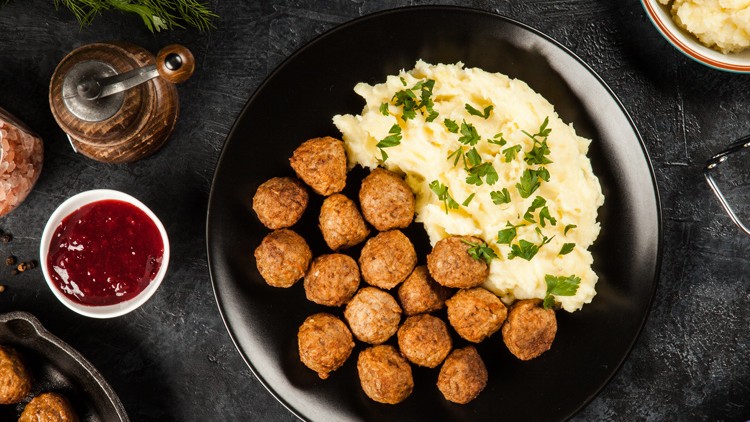 Schwarzer Teller mit Kartoffelpüree, dekoriert mit Kräutern, und gebratenen Fleischbällchen, daneben Preiselbeersauce in Schale.