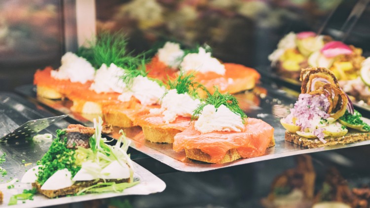 Verschiedene belegte Brote mit Lachs, Kräutern, Frischkäse, Gurken und weiteren Zutaten auf Servierplatten in einer Theke.