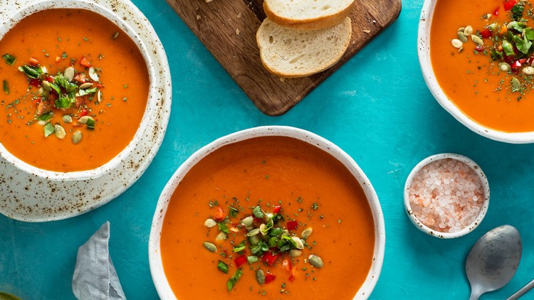 Drei Teller mit roter Suppe, garniert mit Kräutern, Kernen und Gewürzen, daneben Brot, Salz und Besteck auf türkisem Untergrund.