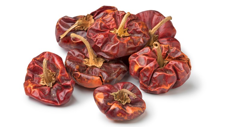 Mehrere kleine, runde, getrocknete Chilischoten mit roten, leicht schrumpeligen Oberflächen und Stielen liegen auf weißem Untergrund.