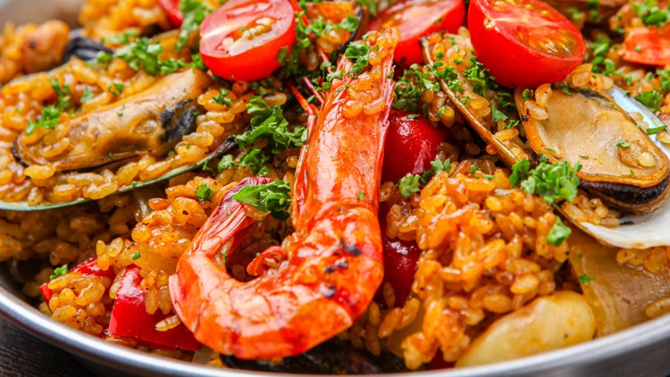 Ein Teller mit Paella, serviert mit Garnelen, Muscheln, roten Paprikastücken, Tomaten und grüner Petersilie.