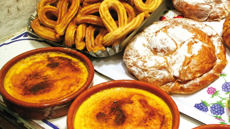 Diverse Backwaren wie Croissant, Churros und Puderzucker-Gebäck sowie zwei Schälchen mit Crème brûlée auf einem Tisch.