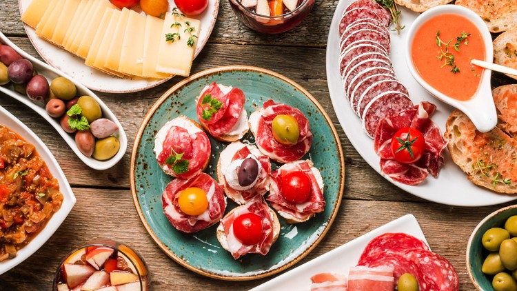 Verschiedene mediterrane Speisen wie Oliven, Salami, Käse, Brot, Aufstrich und Dips auf Holztisch angerichtet.