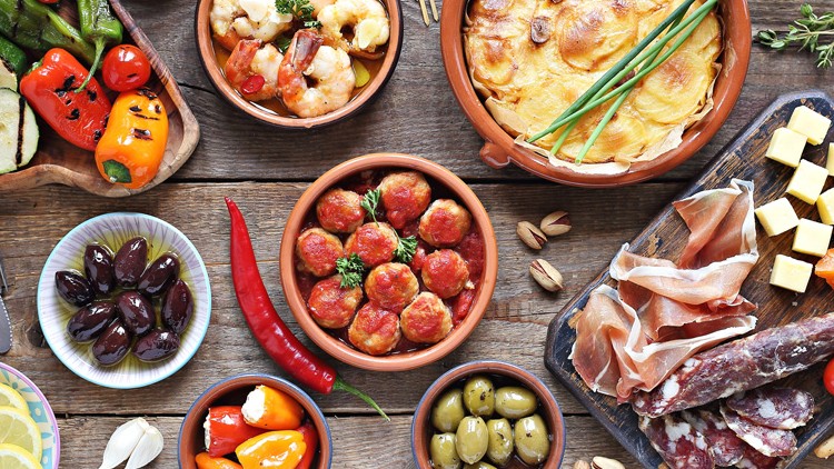 Mediterrane Speisen wie Antipasti, Fleisch, Käsewürfel, Oliven und Auflauf sind auf einem Holztisch angerichtet.