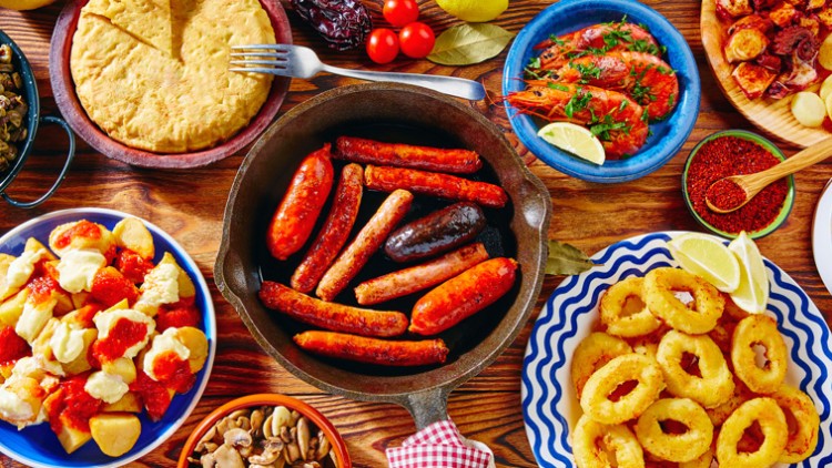 Verschiedene Speisen wie Würste, Garnelen, Kartoffeln, Bohnen, Muscheln, Calamari und Fladenbrot auf einem Holztisch.