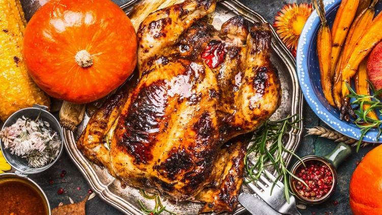 Gebratenes Hähnchen mit Kürbis, gegrilltem Gemüse, Maiskolben, Apfel, Sonnenblumen und Gewürzen auf einem gedeckten Tisch.