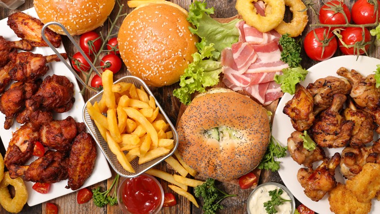 Große Auswahl an Speisen mit Burgern, Pommes, gebratenem Hähnchen, Zwiebelringen, Speck, Salat und Dips auf einem Holztisch.