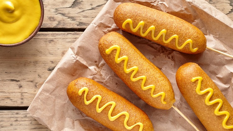 Vier goldbraune Corn Dogs mit gelber Senfdekoration liegen auf braunem Papier, daneben steht eine Schale mit Senf.