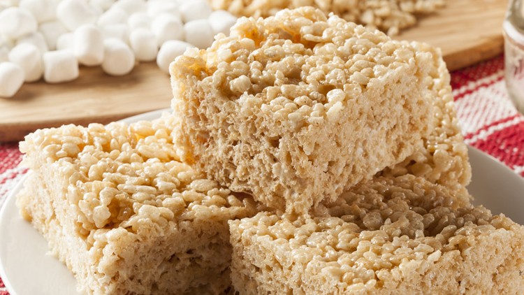 Vier quadratische Reis-Crisp-Riegel auf einem weißen Teller vor Marshmallows und weiteren Riegeln auf Holzbrett.