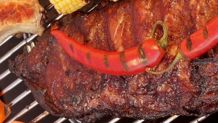 Gegrilltes Steak mit roter Chilischote auf einem Grillrost, umgeben von Maiskolben und weiteren Fleischstücken.