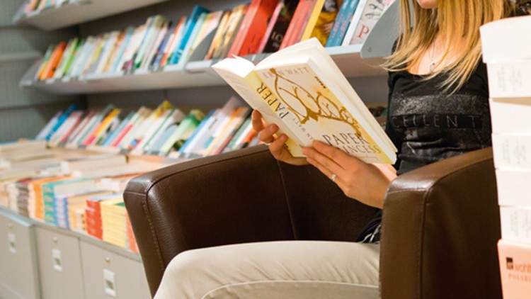 Zwei Personen sitzen auf braunen Sesseln, lesen Bücher, im Hintergrund sind Bücherregale und gestapelte Bücher zu sehen.