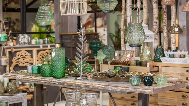 Tisch mit dekorativen Glasgefäßen in Grün- und Brauntönen, Metalltablett, Blattform-Deko und Hängelampen aus Glas und Holz.