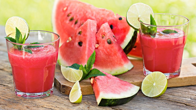 Zwei Gläser Wassermelonensaft mit Minzblättern, Limettenscheiben und frischen Wassermelonenstücken auf einem Holztisch.