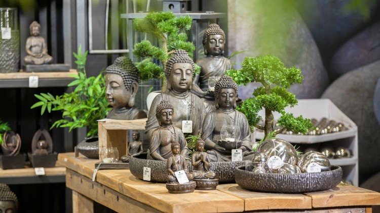 Bronzefarbene Buddha-Statuen, Bonsais und silberne Dekokugeln auf Holztisch, umgeben von Grünpflanzen und Regalen.