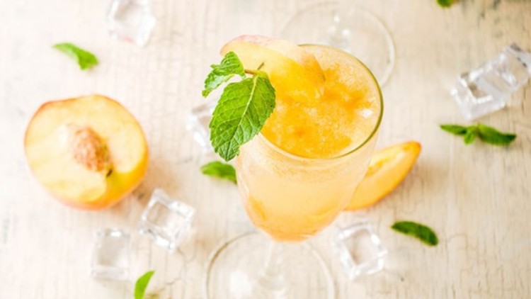 Ein Glas mit pfirsichfarbener Eismischung, garniert mit Minzblatt und Pfirsichspalte, steht auf hellen Holz mit Eiswürfeln und Pfirsichen.