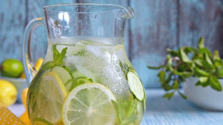 Eine Glaskaraffe mit Wasser, Zitronen-, Gurkenscheiben, Eiswürfeln und Minzblättern steht auf einem gelben Tuch mit weißen Punkten.