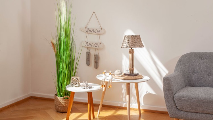 Helles Wohnzimmer mit grauem Sessel, zwei weißen Beistelltischen, Deko-Gegenständen und großer Pflanze in einem Korb.