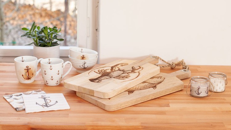 Holztisch mit maritimen Dekorartikeln wie Tassen, Schalen, Schneidebrettern, Servietten und Gläsern, Fenster im Hintergrund.