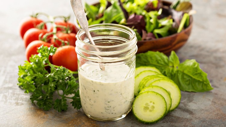 Ein Glas mit cremigem Kräuterdressing, umgeben von Petersilie, Tomaten, Gurkenscheiben, Basilikum und einer Schüssel Salat.