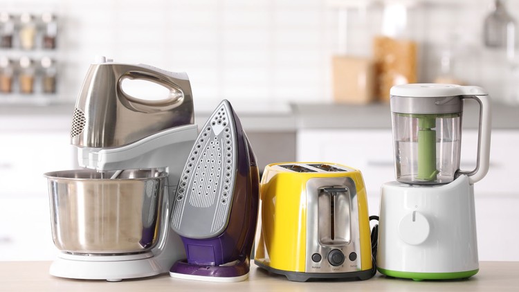 Eine Küchenmaschine, ein Bügeleisen, ein gelber Toaster und ein Mixer stehen nebeneinander auf einer Arbeitsfläche.