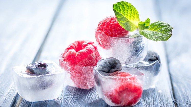 Gefrorene Himbeeren und Blaubeeren in Eiswürfeln, dekoriert mit frischen Minzblättern auf einer hellen Holzoberfläche.