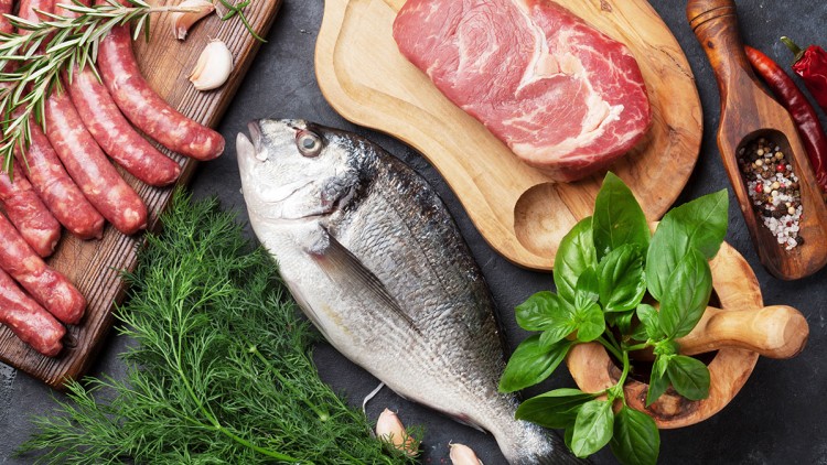 Rohes Rindersteak auf Holzbrett, frischer Fisch, rohe Würste, Kräuter wie Dill und Basilikum sowie Gewürze und Knoblauch.