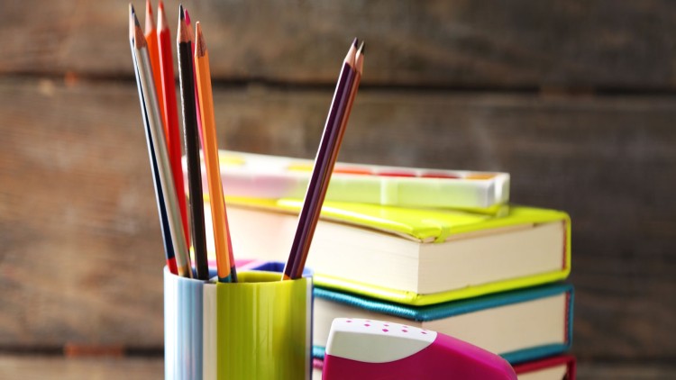 Bunte Stifte in einem Becher, ein Tintenkorrekturroller, Bücher und ein Malkasten auf einem Holztisch.